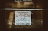 A printed sign is taped to a glass door. The sign contains a message encouraging people to recognize their own white privilege and engage in difficult conversations with family and friends to create change. The background is slightly blurred and appears to be outdoors.