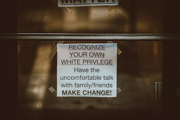 A printed sign is taped to a glass door. The sign contains a message encouraging people to recognize their own white privilege and engage in difficult conversations with family and friends to create change. The background is slightly blurred and appears to be outdoors.