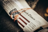 A newspaper is tied with an orange rubber band, featuring a headline and an image of a smiling person alongside an American flag pattern. The text and image are partially visible.