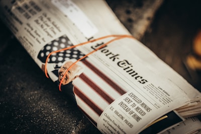 A newspaper is tied with an orange rubber band, featuring a headline and an image of a smiling person alongside an American flag pattern. The text and image are partially visible.