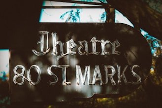 A vintage-style sign reads 'Theatre 80 St Marks' with a retro aesthetic, set against a backdrop of trees that create a dappled sunlight effect.