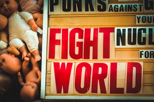 A display combines dolls with a sign bearing bold text conveying a message against nuclear proliferation. The left side of the image is filled with doll parts, some showing faces with open eyes. The right side of the image features a board with large, colorful letters forming words like 'FIGHT' and 'WORLD'.