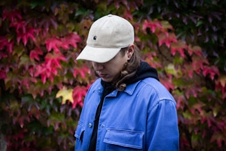 A person stands against a backdrop of vibrant autumn leaves. They are wearing a beige cap and a blue jacket, gazing downward with a contemplative expression. The foliage features hues of red, pink, and green, adding a rich tapestry of colors to the scene.