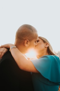 A couple is embracing and kissing, with the sunlight streaming between them. The woman's hand is wrapped around the man's neck, and she is wearing a wristwatch and earrings. The scene conveys intimacy and affection.