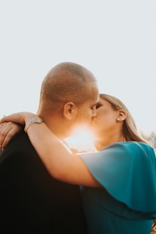 A couple is embracing and kissing, with the sunlight streaming between them. The woman's hand is wrapped around the man's neck, and she is wearing a wristwatch and earrings. The scene conveys intimacy and affection.