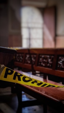 A dimly lit indoor scene featuring wooden benches and a yellow caution tape with the word 'PROHIBIDO' in black letters, indicating restricted access. The background is blurred, with visible outlines of windows and structures typical of a church or gathering hall.
