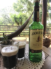 A bottle of Jameson Irish Whiskey is placed on a wooden table with two glasses filled with a dark beverage topped with creamy foam. The scene includes a crocheted table mat, a small succulent plant, and a background of lush greenery and an outdoor hammock. The setting appears relaxed and inviting, suggesting an outdoor gathering.