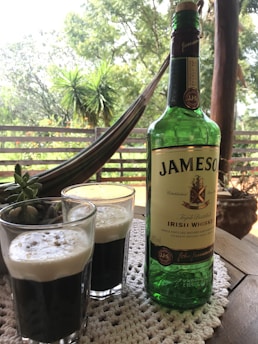 A bottle of Jameson Irish Whiskey is placed on a wooden table with two glasses filled with a dark beverage topped with creamy foam. The scene includes a crocheted table mat, a small succulent plant, and a background of lush greenery and an outdoor hammock. The setting appears relaxed and inviting, suggesting an outdoor gathering.