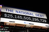 A digital billboard displays the current value of the national debt with bold numerals in white against a black background. Above the numbers, the phrase 'The National Debt' is prominently written in blue on a white section of the sign. The sign appears to be set outdoors at night, illuminated by external lighting.