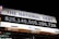 A digital billboard displays the current value of the national debt with bold numerals in white against a black background. Above the numbers, the phrase 'The National Debt' is prominently written in blue on a white section of the sign. The sign appears to be set outdoors at night, illuminated by external lighting.