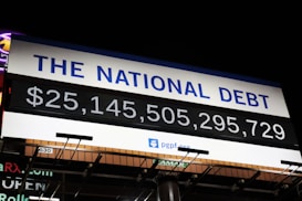 A digital billboard displays the current value of the national debt with bold numerals in white against a black background. Above the numbers, the phrase 'The National Debt' is prominently written in blue on a white section of the sign. The sign appears to be set outdoors at night, illuminated by external lighting.