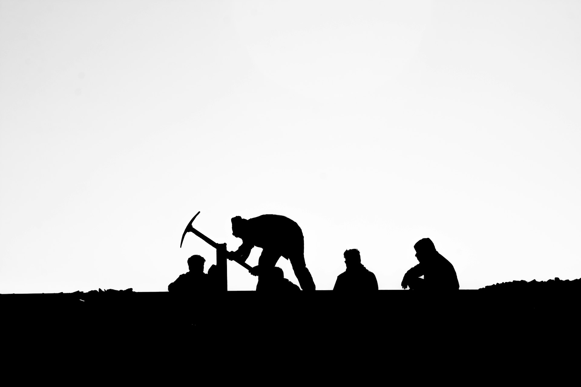 Silhouettes of laborers with a pickaxe against a bright sky, high contrast.