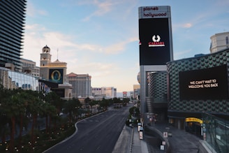 A view of a wide, empty city street lined with palm trees and modern buildings. Prominent buildings include a recognizable tower with a large digital sign displaying 'Caesars Entertainment' and another message 'We can't wait to welcome you back'. The street is well-maintained and devoid of traffic. The skyline features a mix of architecture styles with a clear blue sky.