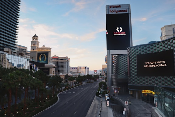 A view of a wide, empty city street lined with palm trees and modern buildings. Prominent buildings include a recognizable tower with a large digital sign displaying 'Caesars Entertainment' and another message 'We can't wait to welcome you back'. The street is well-maintained and devoid of traffic. The skyline features a mix of architecture styles with a clear blue sky.