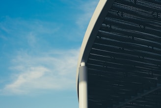 A large sectional view of a metal structure appears to extend towards the sky, with its surface featuring a series of small perforations. The structure is supported by a pillar, and the background consists of a clear blue sky with some scattered white clouds.