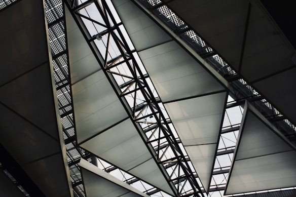 A geometric, metal and glass architectural structure featuring intersecting lines and triangular panels. The panels allow natural light to filter through, creating a dynamic and modern design.