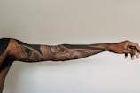 An extended arm featuring intricate black ink tattoos with geometric and abstract designs. The tattoos cover the entire visible length of the arm, which is set against a neutral, light background.