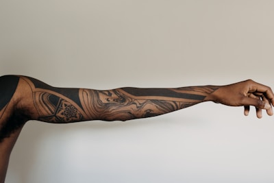 An extended arm featuring intricate black ink tattoos with geometric and abstract designs. The tattoos cover the entire visible length of the arm, which is set against a neutral, light background.