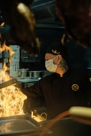A chef wearing a black uniform and a face mask works intensely in a dimly lit kitchen with flames rising from a stovetop. The environment suggests a professional setting with various kitchen utensils and equipment in the background.