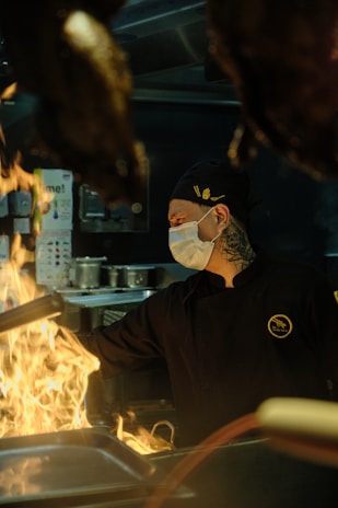A chef wearing a black uniform and a face mask works intensely in a dimly lit kitchen with flames rising from a stovetop. The environment suggests a professional setting with various kitchen utensils and equipment in the background.
