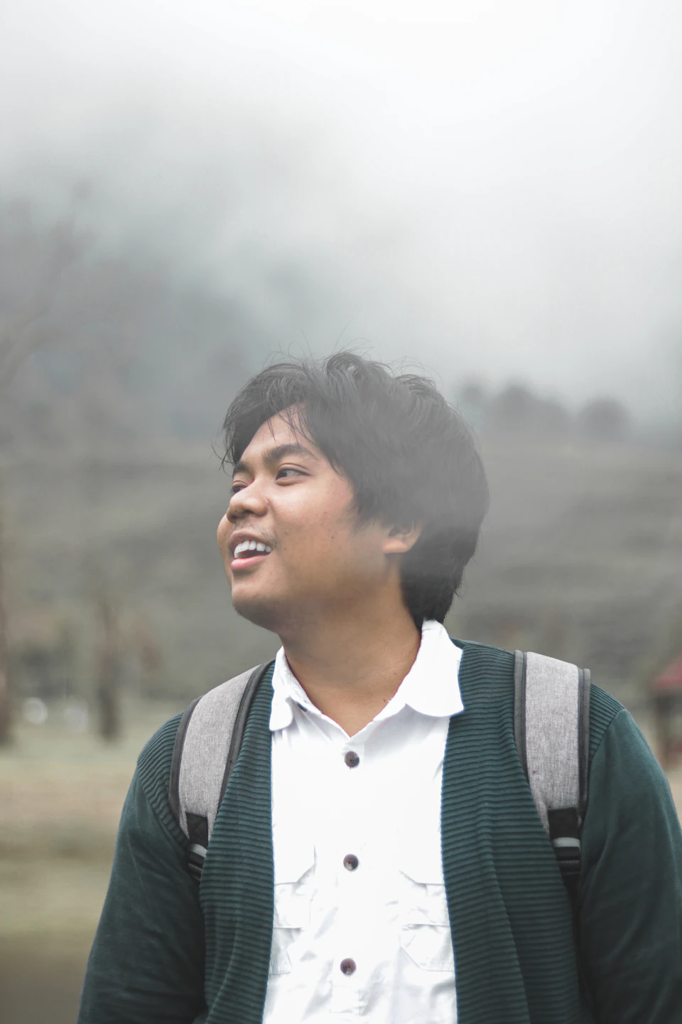 In the image, a young man with dark hair is standing against a backdrop of foggy mountains and trees. He's dressed casually for the day, wearing a white shirt and a green sweater vest over it. A backpack rests on his shoulders, suggesting he may be out exploring or hiking in the area. The man has a warm smile on his face as he looks up at something above him, possibly admiring the view of the foggy landscape or perhaps spotting someone else in the distance.

The image is taken from an angle that places the man centrally and slightly to the left, giving prominence to both the subject and the surrounding environment. The foggy mountains and trees in the background add a sense of depth and mystery to the scene, while the man's bright smile and casual attire create a warm and inviting atmosphere.