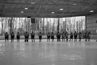 A lineup of ice hockey players stands in an indoor rink, facing away and wearing jerseys with numbers. An American flag hangs prominently in the background, with trees visible through large windows.