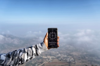 A person extends an arm clad in a patterned jacket, holding a smartphone displaying a compass app. The background features a landscape with mist and clouds, creating a serene, expansive view from a high vantage point.