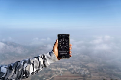 A person extends an arm clad in a patterned jacket, holding a smartphone displaying a compass app. The background features a landscape with mist and clouds, creating a serene, expansive view from a high vantage point.