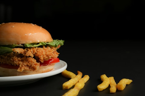 A crispy fried chicken sandwich with lettuce and tomato is placed on a white plate. Next to it, a few curly fries are scattered on a dark surface, creating a simple yet appetizing presentation.