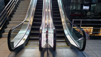 An escalator and staircase are positioned side by side in a public indoor environment. The escalator has two sets of moving stairs with shiny metal railings, and there are visible warning signs posted at the foot. The surrounding area includes tiled flooring and metal railings, and part of a building interior with glass and furniture can be seen in the background.
