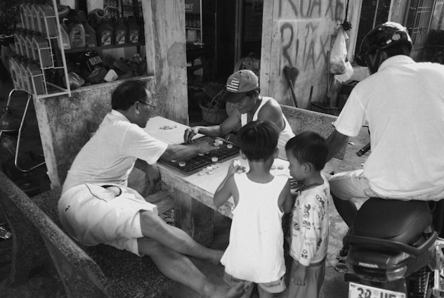 Several people are gathered around an outdoor stone table, engaged in a board game. Two adults are seated, intensely focused on the game, while a third adult observes from a motorbike. Two children stand nearby, watching the game with curiosity. The setting appears casual, with a backdrop of a simple storefront and supplies nearby.
