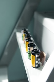 Several rolls of camera film are lined up on a narrow white shelf. The film rolls have different labels with black, yellow, and green colors. One of the rolls has a portion of film partially unrolled, exposing the brown film strip.