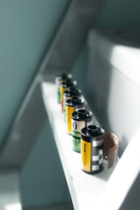 Several rolls of camera film are lined up on a narrow white shelf. The film rolls have different labels with black, yellow, and green colors. One of the rolls has a portion of film partially unrolled, exposing the brown film strip.