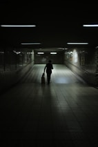 A dimly lit underground passageway with tiled walls and floor, illuminated by fluorescent lights. A solitary figure walks away, silhouetted against the light, pulling a small suitcase. The atmosphere is quiet and contemplative.