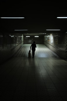 A dimly lit underground passageway with tiled walls and floor, illuminated by fluorescent lights. A solitary figure walks away, silhouetted against the light, pulling a small suitcase. The atmosphere is quiet and contemplative.