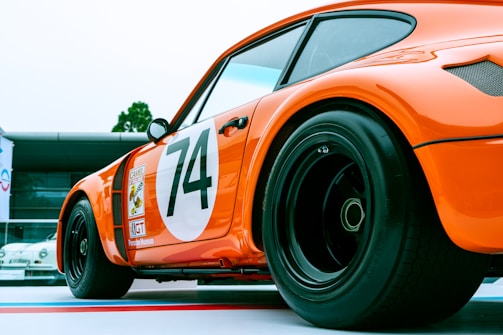 A bright orange vintage racing car with a large number 74 on its side is prominently displayed. The car has sleek, aerodynamic lines and wide, black racing tires. The background includes a modern building with glass elements, contrasting with the classic design of the car.