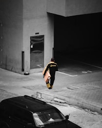A delivery person wearing a black and yellow uniform is walking with a parcel in hand. The scene appears to be in an urban area, near a building entrance, with a parked vehicle visible in the foreground. The environment looks subdued and industrial, with concrete surfaces and muted tones.