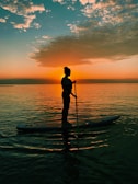 A person is paddleboarding on calm water during a vibrant sunset. The sky is filled with a mix of orange and blue hues, with clouds scattered across the sky. The silhouette of the person contrasts against the fiery horizon, creating a serene and picturesque scene.