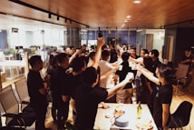 A group of people stand around a table in an office setting, raising glasses in a toast. The room features glass walls, office chairs, and a wooden ceiling. The table is adorned with a variety of snacks and a few bottles. The gathering appears to be a celebratory or social event.