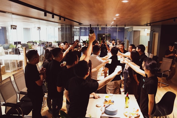 A group of people stand around a table in an office setting, raising glasses in a toast. The room features glass walls, office chairs, and a wooden ceiling. The table is adorned with a variety of snacks and a few bottles. The gathering appears to be a celebratory or social event.