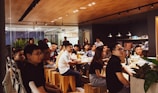 A group of people is gathered in a modern, well-lit office space with a wooden ceiling and glass walls. They are sitting on wooden stools around a long table, attentively listening or watching something. There are bottles and snacks on the table, and the space is decorated with plants.