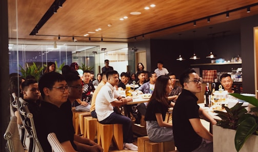 A group of people is gathered in a modern, well-lit office space with a wooden ceiling and glass walls. They are sitting on wooden stools around a long table, attentively listening or watching something. There are bottles and snacks on the table, and the space is decorated with plants.