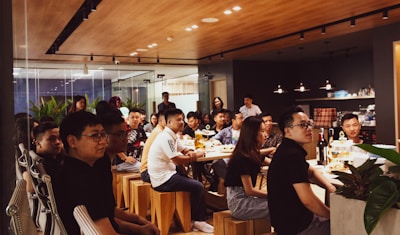 A group of people is gathered in a modern, well-lit office space with a wooden ceiling and glass walls. They are sitting on wooden stools around a long table, attentively listening or watching something. There are bottles and snacks on the table, and the space is decorated with plants.