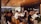 A group of people is gathered in a modern, well-lit office space with a wooden ceiling and glass walls. They are sitting on wooden stools around a long table, attentively listening or watching something. There are bottles and snacks on the table, and the space is decorated with plants.
