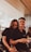 A warm and friendly scene featuring two people sitting together in an office or meeting space. They both wear matching black shirts, with one person wearing glasses. The setting includes glass walls, modern office chairs, and other individuals blurred in the background.