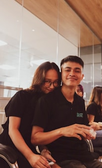 A warm and friendly scene featuring two people sitting together in an office or meeting space. They both wear matching black shirts, with one person wearing glasses. The setting includes glass walls, modern office chairs, and other individuals blurred in the background.