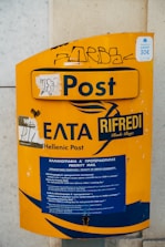 A yellow postal box with the label 'Hellenic Post' and 'Post' prominently displayed. It features some graffiti and stickers, including a sign indicating a 30 euro airport transfer. The box contains a section for priority mail, with information in both Greek and English.