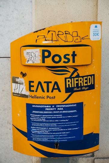 A yellow postal box with the label 'Hellenic Post' and 'Post' prominently displayed. It features some graffiti and stickers, including a sign indicating a 30 euro airport transfer. The box contains a section for priority mail, with information in both Greek and English.