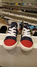 A display features a pair of children's sneakers with navy blue fabric and bright red toes, white laces, and white soles. They are placed on a shelf in a shoe store with other footwear visible in the background, arranged neatly on several shelves.