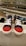 A display features a pair of children's sneakers with navy blue fabric and bright red toes, white laces, and white soles. They are placed on a shelf in a shoe store with other footwear visible in the background, arranged neatly on several shelves.